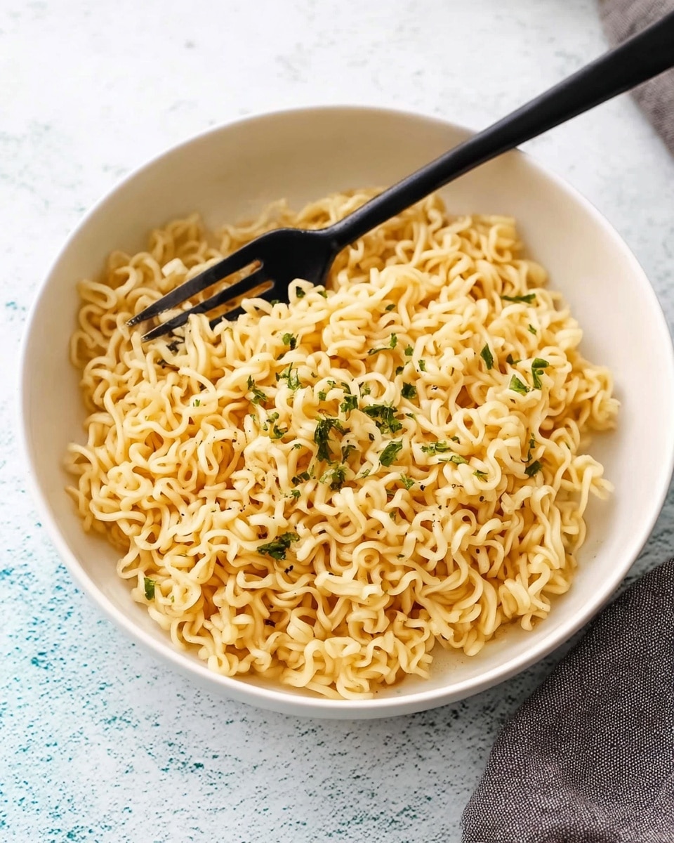 A white bowl filled with cooked, light yellow instant noodles topped with small green herb pieces, giving a fresh contrast. A black fork rests inside the bowl, its prongs tangled lightly in the noodles. The bowl is set against a white marbled textured surface with light blue and white tones visible around it, and part of a gray cloth is seen at the bottom right corner. photo taken with an iphone --ar 4:5 --v 7