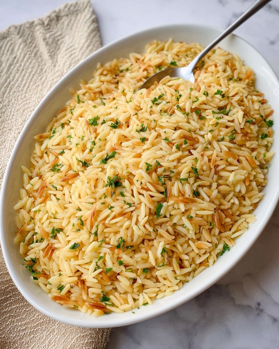 A white oval dish filled with a single layer of cooked rice mixed with orzo, light yellow to golden in color, with some toasted orzo pieces showing a darker brown shade. Small green herb pieces are sprinkled evenly on top, adding a fresh touch. A metal spoon is placed inside the dish, resting on the upper edge. The dish sits on a white marbled surface with a beige textured cloth partially visible on the left side. photo taken with an iphone --ar 4:5 --v 7
