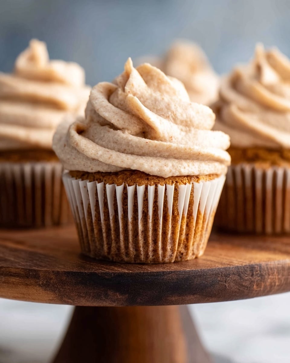 The image shows a close-up of five cupcakes arranged in a soft focus on a white marbled surface. Each cupcake has one layer of golden brown cake with a slightly rough texture, visible through the light brown paper liners. On top of each cupcake is a thick swirl of creamy frosting in a light beige color, speckled with tiny darker dots suggesting cinnamon or nutmeg. The frosting has a smooth, soft texture with defined ridges from piping, creating a tall, conical shape with a small peak at the top. In the foreground, a whole nutmeg seed and a cinnamon stick lay on the surface, adding a rustic, warm touch to the scene. The photo is bright with soft shadows and a clean, minimal background. photo taken with an iphone --ar 4:5 --v 7