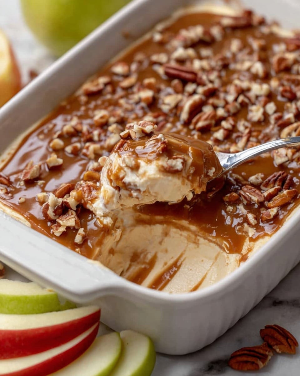 A white rectangular ceramic dish filled with a creamy, light beige dip topped with a thick layer of dark brown caramel sauce and scattered with chopped pecans. A spoon is scooping out a portion of the creamy dip from the corner of the dish, blending the smooth beige and caramel layers together. In the background, fresh green apple slices and red apple slices lean against the side of the dish. The dish is placed on a white marbled surface. photo taken with an iphone --ar 4:5 --v 7