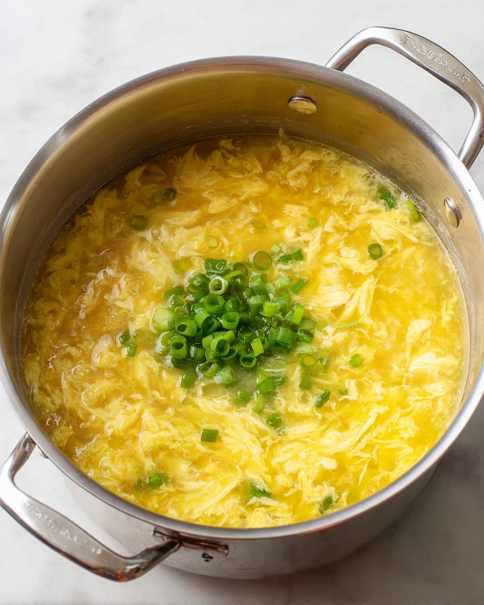A stainless steel pot filled with a thick, yellow egg drop soup that has a soft and silky texture with visible strands of cooked egg swirled throughout. In the center of the soup is a small mound of chopped bright green scallions, adding a fresh contrast in color. The pot has two handles and is set on a white marbled surface. photo taken with an iphone --ar 4:5 --v 7
