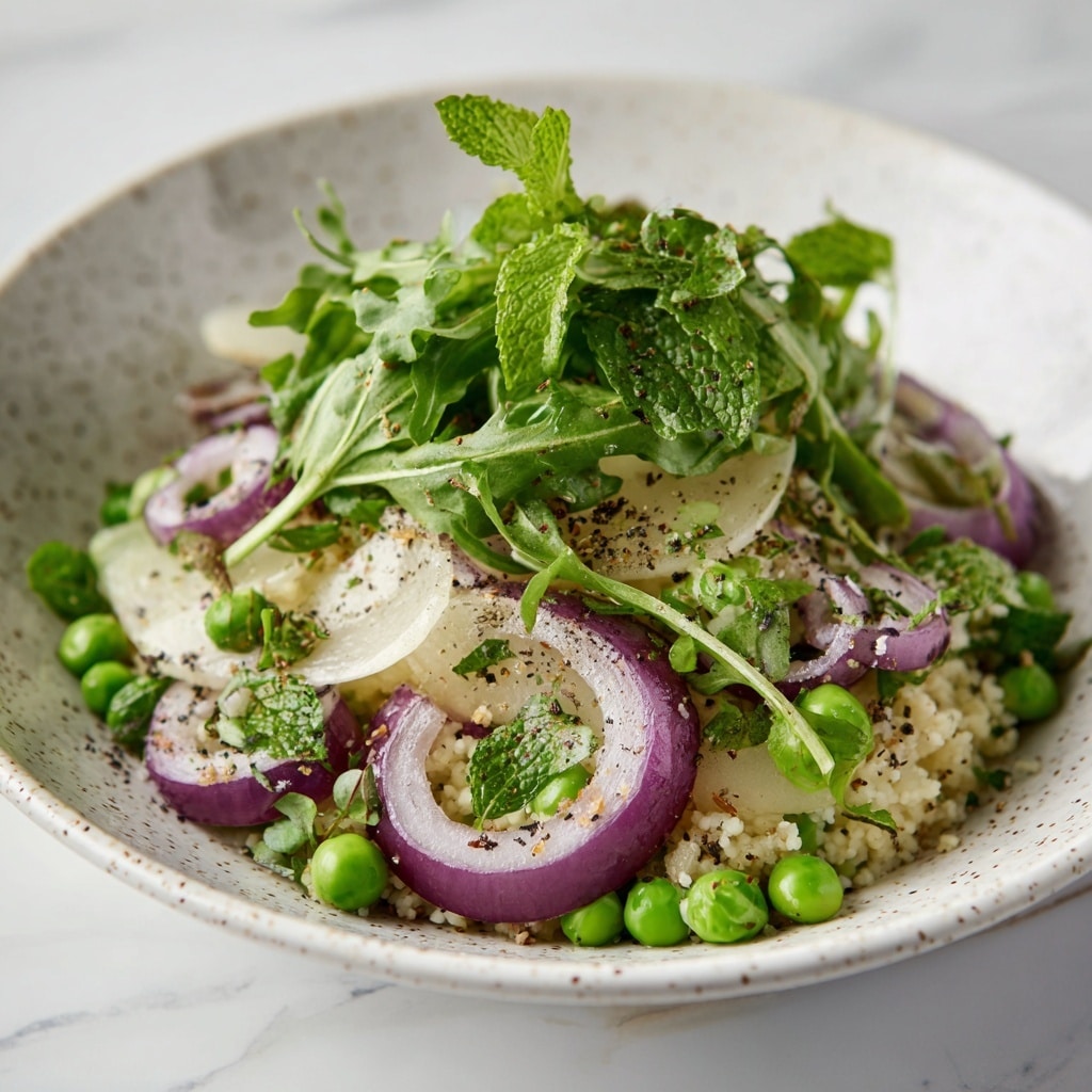 The dish is a fresh salad in a white speckled round bowl, filled with small white couscous pearls scattered throughout. Bright green peas and leafy greens, including arugula and mint leaves, add vibrant green color and soft texture. Thin slices of purple-red onion are layered on top, offering a crisp contrast. The salad is lightly sprinkled with black pepper and herbs, giving a speckled effect over the mix. The bowl sits on a white marbled surface, and the colors of the salad pop against the neutral background. Photo taken with an iphone --ar 4:5 --v 7