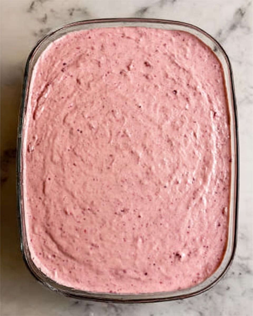 The image shows a white plate with one piece of strawberry cake with three layers: the bottom layer is a pink moist cake, the middle layer is a thick white cream with bits of strawberries, and the top layer is fluffy white whipped cream decorated with strawberry pieces and a whole strawberry on top. A metal fork is cutting into the cake, held by a woman's hand, with bits of cake stuck on the fork. The plate is set on a white marbled textured surface with a soft pink lace cloth partly visible beside it. Photo taken with an iphone --ar 4:5 --v 7