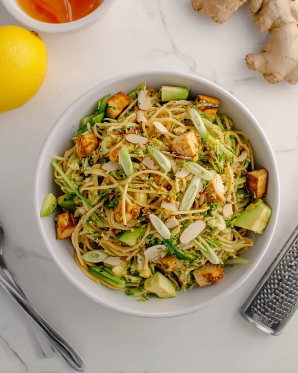 A white bowl filled with a mixed salad sits on a white marbled surface. The salad has several layers starting with thin, light yellow noodles at the base and scattered atop are chunks of golden brown grilled tofu. Bright green sliced scallions and avocado pieces are mixed in, along with thinly sliced almonds adding texture. The salad looks fresh and lightly tossed with visible herb flakes. Nearby on the surface are half a bright yellow lemon, part of ginger root, a metal grater, and a silver fork. photo taken with an iphone --ar 4:5 --v 7