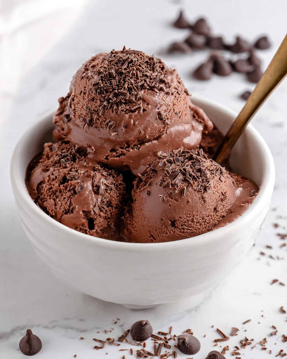 A white bowl filled with three scoops of rich, creamy chocolate ice cream, each scoop smooth but slightly melting at the edges. The ice cream is topped with fine, dark chocolate shavings spread evenly over the surface, adding a textured contrast to the smoothness. Around the bowl, there are scattered chocolate chips and small chocolate shavings on a white marbled surface. A small bronze spoon is partially visible inside the bowl, resting against the side. The photo taken with an iphone --ar 4:5 --v 7