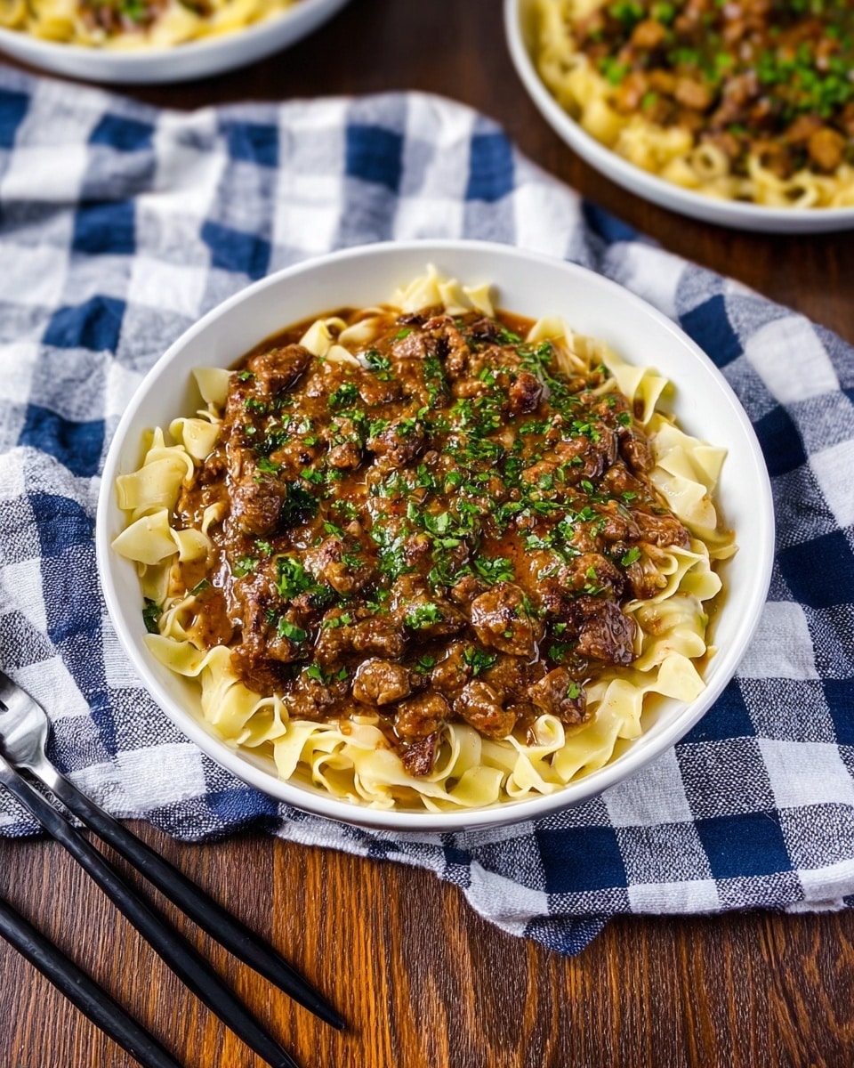 A white bowl filled with three layers of food: the bottom layer is wide, flat, pale yellow noodles; the middle layer is a thick, brown sauce with small pieces of cooked meat mixed in; the top layer is finely chopped green herbs sprinkled evenly over the brown sauce and meat. The bowl is placed on a dark brown wooden surface, surrounded by a white and blue striped and checkered cloth, and there are black chopsticks and two forks positioned to the left side of the bowl, with two smaller dishes filled with the same food visible in the background. photo taken with an iphone --ar 4:5 --v 7