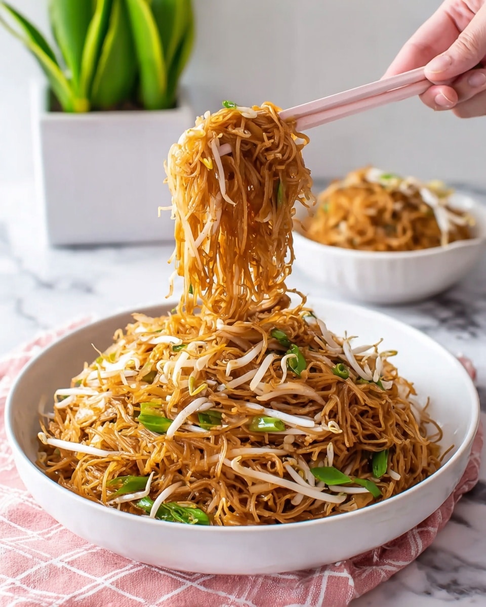 A white round bowl is filled with a large pile of stir-fried noodles that have a shiny, dark orange-brown color. Mixed into the noodles are thin white bean sprouts and pieces of green leafy vegetables scattered throughout, adding contrast. A woman's hand holds pale pink chopsticks, lifting a clump of noodles high above the bowl, showing the long, tangled strings falling down. In the background, there is a second white bowl with more noodles and a green plant in a white planter sitting on a white marbled surface. The scene is softly lit with natural light, creating a fresh and inviting feel. photo taken with an iphone --ar 4:5 --v 7