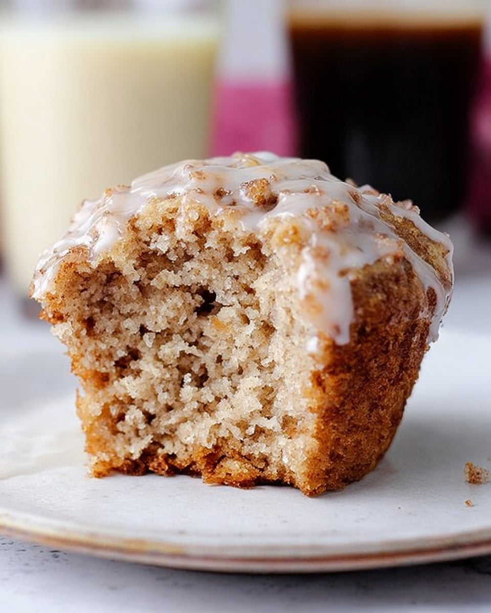 A single cinnamon muffin sits on a small white plate, showing a rough, crumbly top layer with bits of cinnamon strewn throughout; the muffin is drizzled with a thin white glaze that flows down the sides in small streams. The plate rests on a bright pink floral napkin placed on a white marbled surface. A silver fork lies on the left side of the plate, its shiny texture catching the light. The background softly blurs out glass jars or cups, adding depth to the image. Photo taken with an iphone --ar 4:5 --v 7