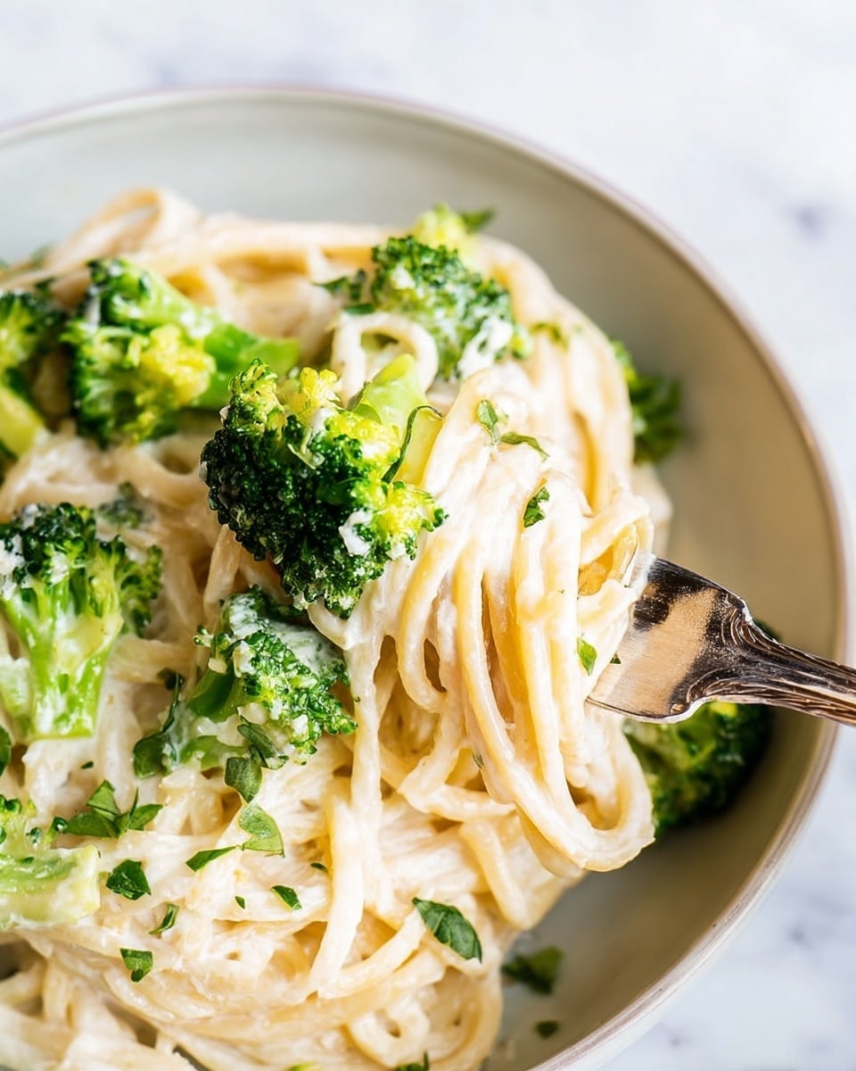 A close-up of a white bowl filled with creamy spaghetti pasta, coated in a smooth, light beige sauce with a slightly glossy texture. The pasta strands are tangled and piled up in the bowl, with a woman's hand holding a silver fork twirling a small bundle of spaghetti near the center. The dish is sprinkled with small bits of chopped green herbs scattered on top, adding contrast to the creamy sauce. The bowl sits on a white marbled surface, and a blurred glass with a golden liquid is visible in the background. photo taken with an iphone --ar 4:5 --v 7