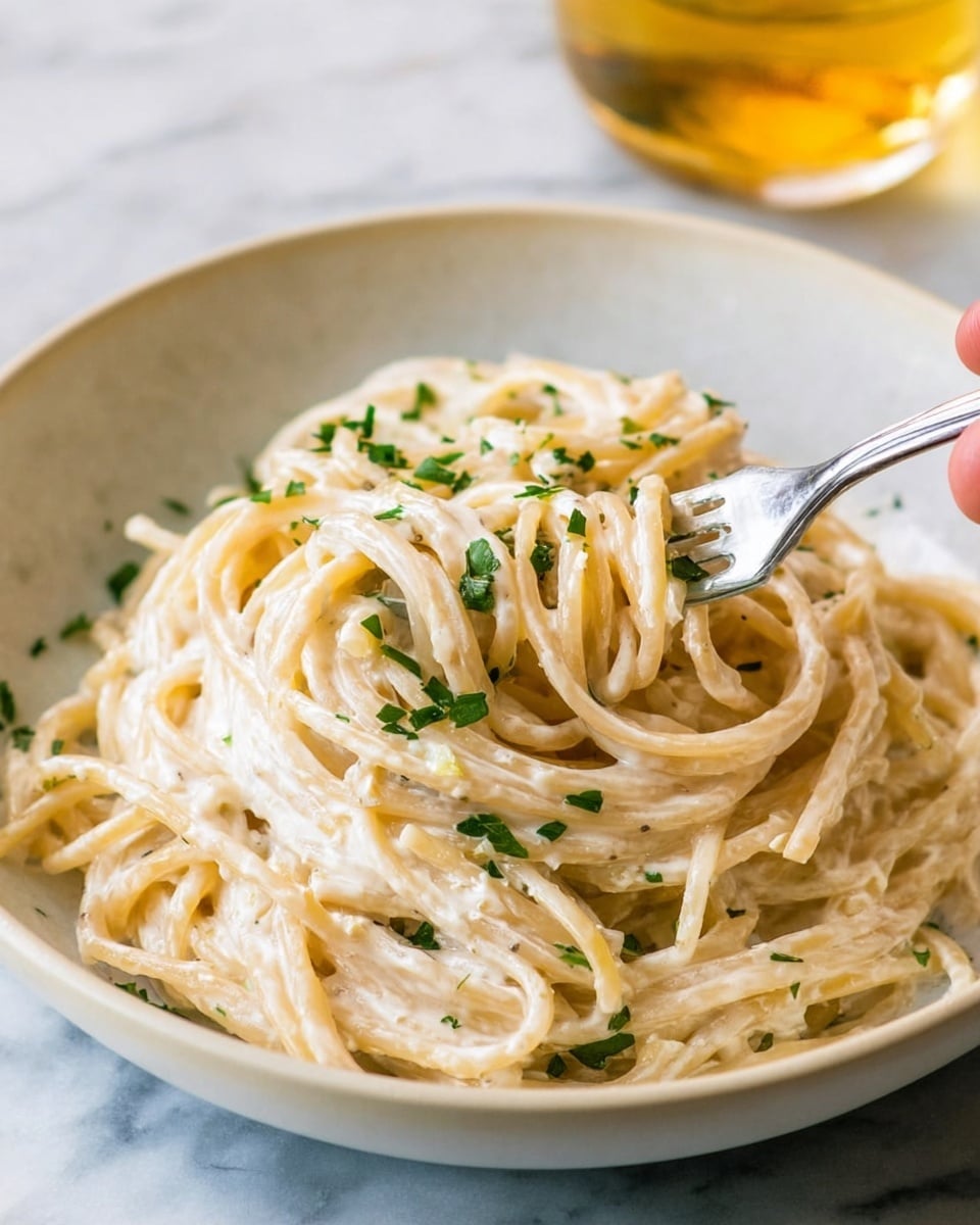 A close-up of creamy spaghetti pasta coated in a smooth white sauce, with bright green broccoli florets mixed in and scattered on top. The pasta strands are thick and twisted around the prongs of a shiny fork held from the right side of the image. Small bits of fresh green herbs are sprinkled on the pasta and broccoli, adding texture and color. The food is served in a simple white bowl, placed on a white marbled surface. photo taken with an iphone --ar 4:5 --v 7
