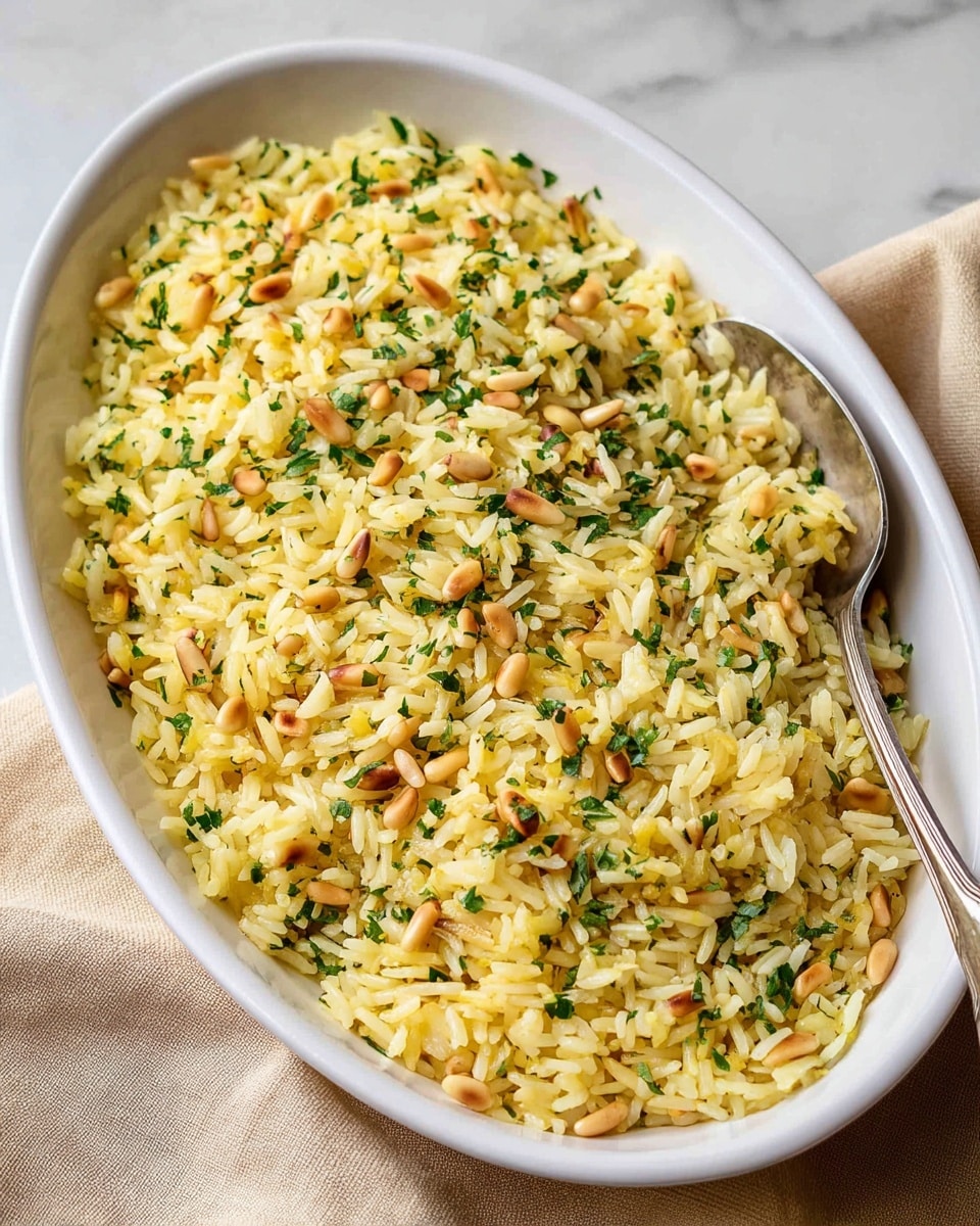 A white oval dish filled with one layer of yellow cooked rice mixed with small toasted pine nuts and chopped green herbs evenly spread throughout. A silver spoon rests partly inside the rice on the right side. The dish is placed on a white marbled surface with a beige cloth partially visible in the background. photo taken with an iphone --ar 4:5 --v 7