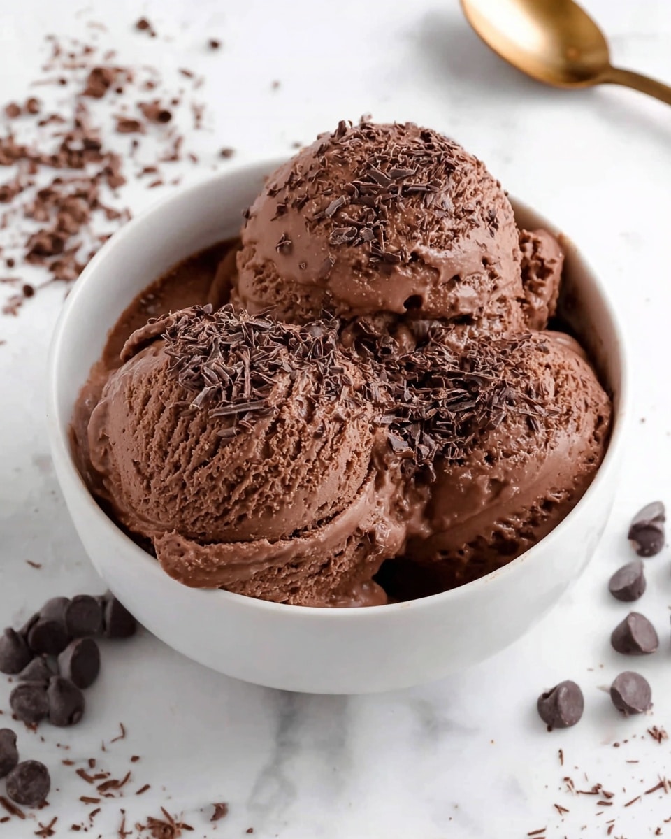 A white ceramic bowl filled with three scoops of rich, creamy dark chocolate ice cream, each scoop smooth and slightly melting at the edges. The ice cream is sprinkled generously with finely grated chocolate shavings on top, adding a textured layer of dark brown specks. A bronze spoon is inserted into the right side of the bowl. Around the bowl on the white marbled surface, there are scattered semi-melted chocolate chips and chocolate flakes, adding a playful touch. The photo taken with an iphone --ar 4:5 --v 7