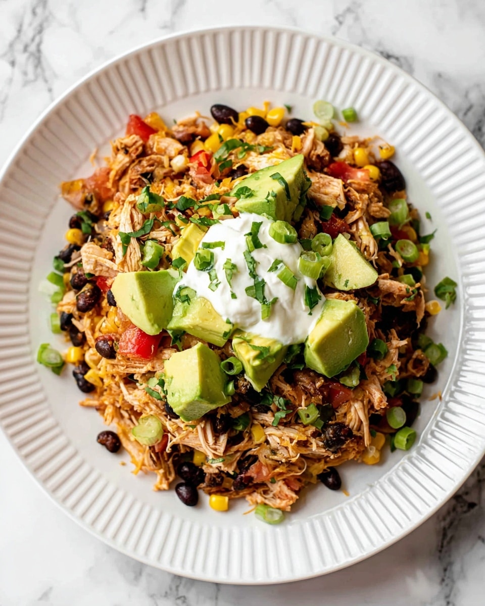 A white ridged plate holds a colorful mixed dish with four visible layers: a base layer of shredded light brown chicken mixed with black beans, yellow corn, and small red pepper pieces; scattered small green onion slices and fresh chopped herbs on top; vibrant green chunks of avocado placed evenly around and on the chicken mixture; and a dollop of white creamy sauce sitting in the center. The plate rests on a white marbled surface. photo taken with an iphone --ar 4:5 --v 7