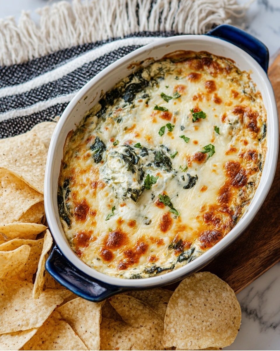 A white oval baking dish filled with a baked spinach artichoke dip, showing one thick layer with a creamy white base mixed with chopped dark green spinach and pale yellow artichoke pieces, topped with a melted golden-brown cheese crust and scattered small green parsley pieces. The dish is placed on a white marbled surface, next to a pile of light-colored tortilla chips with a rough texture and small brown toasted spots. photo taken with an iphone --ar 4:5 --v 7
