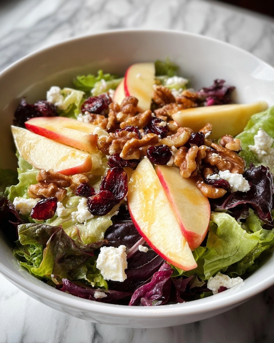 A fresh salad in a white bowl consists of one layer of mixed green and purple lettuce leaves as the base. Above the greens, there are several triangular slices of red-skinned apple with pale yellow flesh. Scattered on top are small white crumbles of soft cheese and dark red dried cranberries. The salad is finished with a generous layer of whole walnut halves that have a glossy texture, likely from a light dressing. The bowl sits on a white marbled surface. Photo taken with an iphone --ar 4:5 --v 7