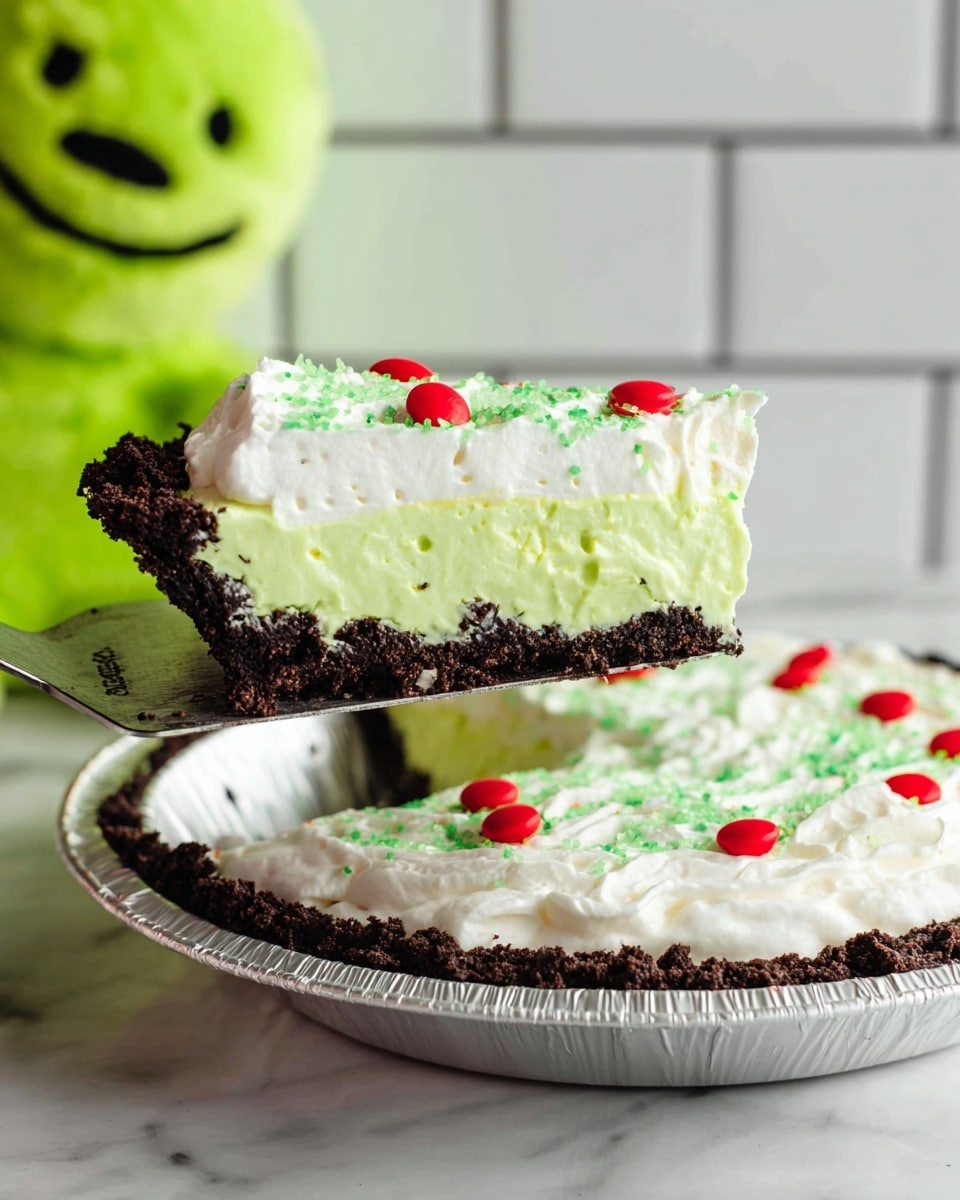 A slice of pie is held on a metal pie server above a round white foil pie dish. The pie has three layers: a dark crumbly cookie crust at the bottom, a thick bright green creamy filling in the middle, and a smooth white whipped cream layer on top. Bright green sprinkles lightly cover the white layer, with small red candy pieces evenly scattered on top. The background is a white marbled texture with a white tile wall behind. A green plush toy with a black stitched smile is partially visible to the left. photo taken with an iphone --ar 4:5 --v 7