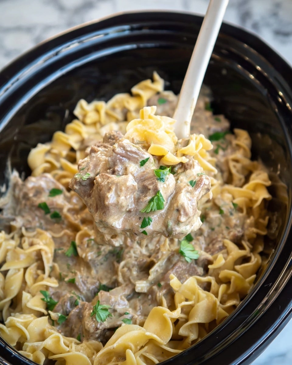 A close-up view of a black slow cooker filled with creamy beef stroganoff, showing three main layers: the bottom layer is light beige egg noodles with a smooth, soft texture, the middle layer consists of chunks of tender beef covered in a thick beige sauce, and the top layer is sprinkled with bright green parsley leaves scattered evenly across the dish. A wooden spoon with a white silicone head rests inside the cooker, its handle sticking out from the top center. The photo is set against a white marbled surface. photo taken with an iphone --ar 4:5 --v 7