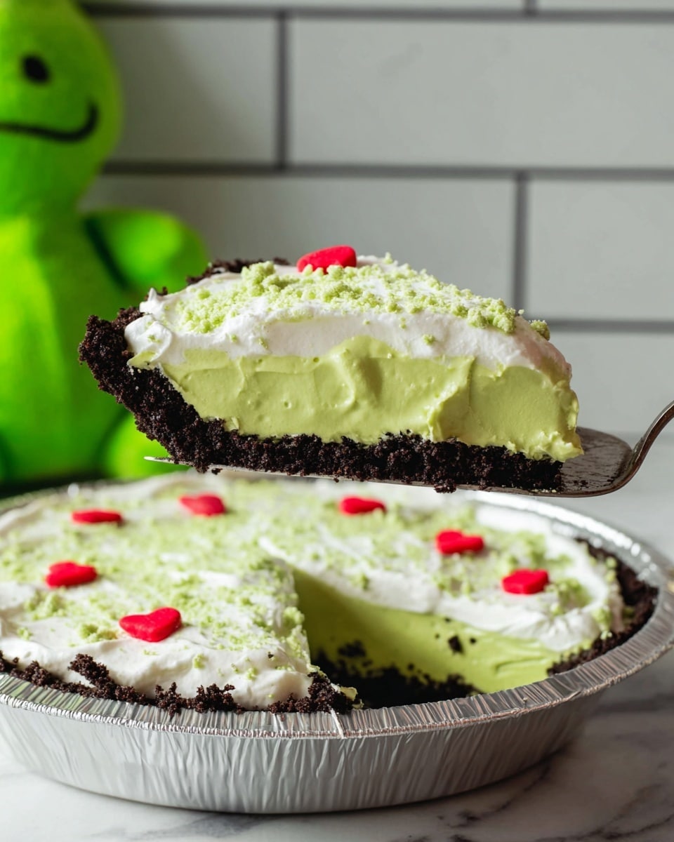 A slice of pie with three visible layers is held above the pie tin by a silver pie server; the bottom layer is a dark crumbly crust, the middle layer is a smooth, thick, bright green filling, and the top layer is a white creamy topping sprinkled with fine green crumbs and dotted with small red heart-shaped decorations. The pie is in a round silver foil pie tin on a white marbled surface with a white tile wall in the background. A green plush toy is partially visible to the left. Photo taken with an iphone --ar 4:5 --v 7