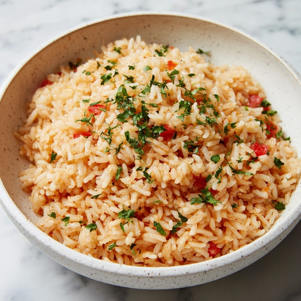 A close-up view of a bowl filled with cooked rice mixed with small chunks of red tomatoes and sprinkled with fresh green herbs on top. The rice looks soft and slightly sticky with a light orange color, and the tomatoes add bright red spots throughout the dish. The bowl is white with a subtle speckled pattern, sitting on a white marbled surface. Photo taken with an iphone --ar 4:5 --v 7