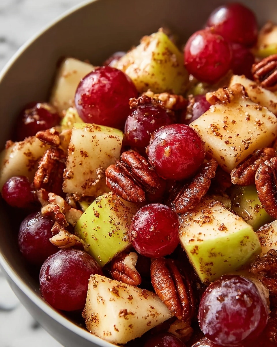 A close-up image of a fruit and nut mix showing round red grapes, small pale yellow apple chunks with a light green tint, and glossy pecans, all sprinkled with a fine brown spice powder. The ingredients are piled thickly together in a white bowl with a smooth texture, placed on a white marbled surface that adds a soft contrast. The grapes are shiny and smooth, the apple pieces have a slightly grainy texture with the spice dusted on them, and the pecans add a crunchy texture with their detailed ridges. photo taken with an iphone --ar 4:5 --v 7