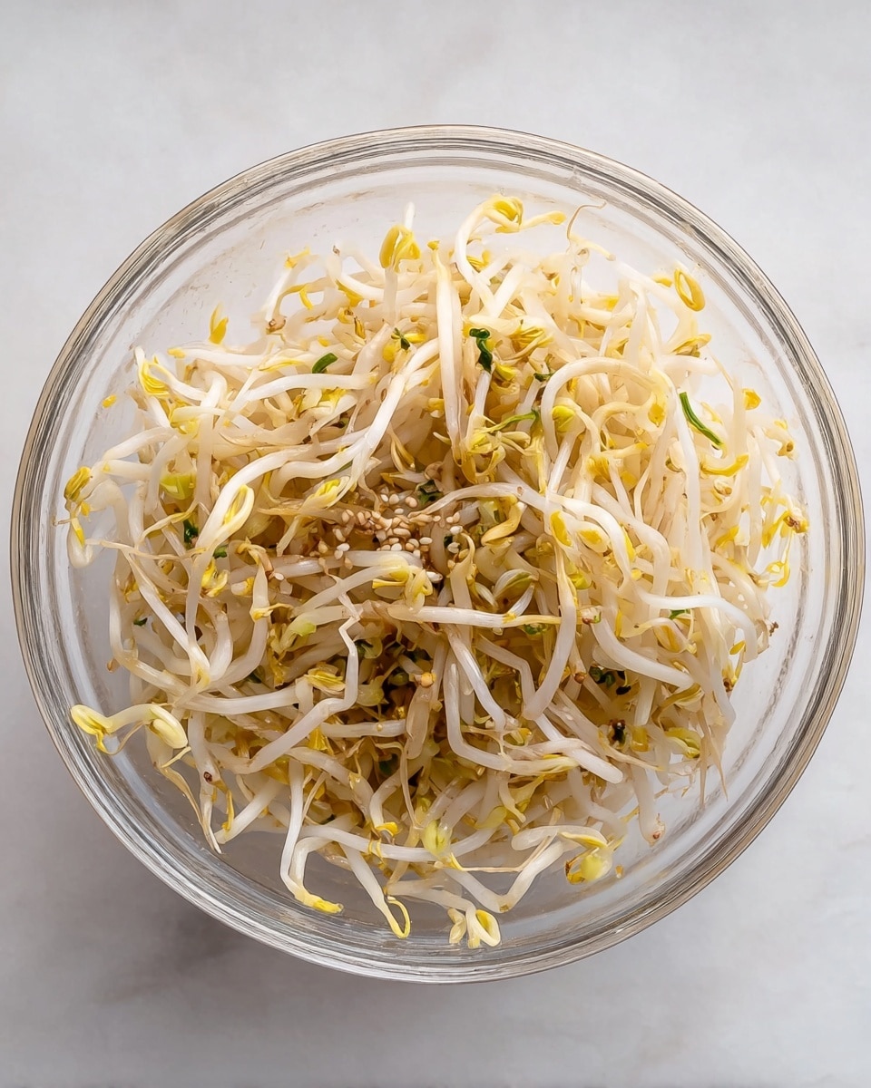 A clear round bowl filled with one layer of lightly cooked soybean sprouts, showing thin, slightly transparent white stems with small yellow tips scattered throughout, mixed with tiny bits of green herbs and sesame seeds. The bowl is placed on a white marbled surface. photo taken with an iphone --ar 4:5 --v 7