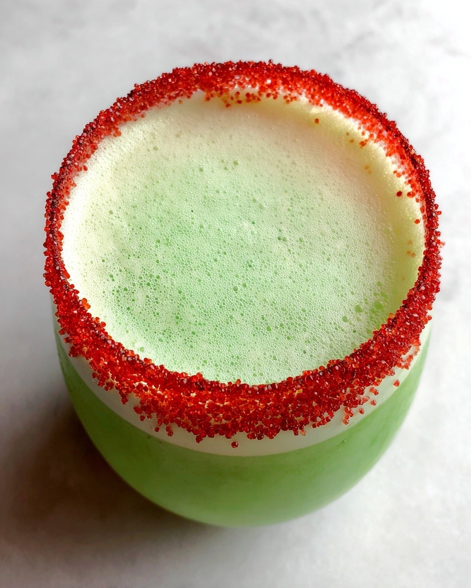A clear glass pitcher with a cartoon green face featuring yellow eyes and a big smile is filled with bright green frothy liquid, topped with a thin layer of white foam. In front of it are two rounded white glasses filled with the same bright green liquid, each with a thick red sugar rim made of coarse granules. The drinks are placed on a clean white marbled surface with a simple white background. photo taken with an iphone --ar 4:5 --v 7