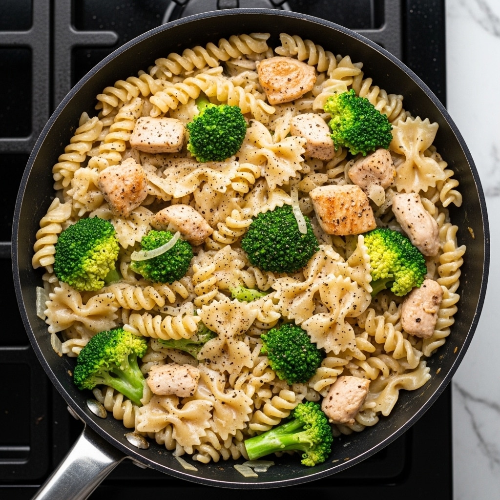 A close-up top view of a black frying pan filled with a creamy pasta dish showing three types of pasta: fusilli, farfalle, and rotini, all coated in a light cream sauce. Scattered throughout are bright green broccoli florets adding a fresh pop of color, along with light brown chunks of cooked chicken breast. The dish is sprinkled with black pepper, giving a contrast to the creamy and green colors. The pan is set on a stove with a dark background, though the surface is imagined as white marbled texture. Photo taken with an iphone --ar 4:5 --v 7
