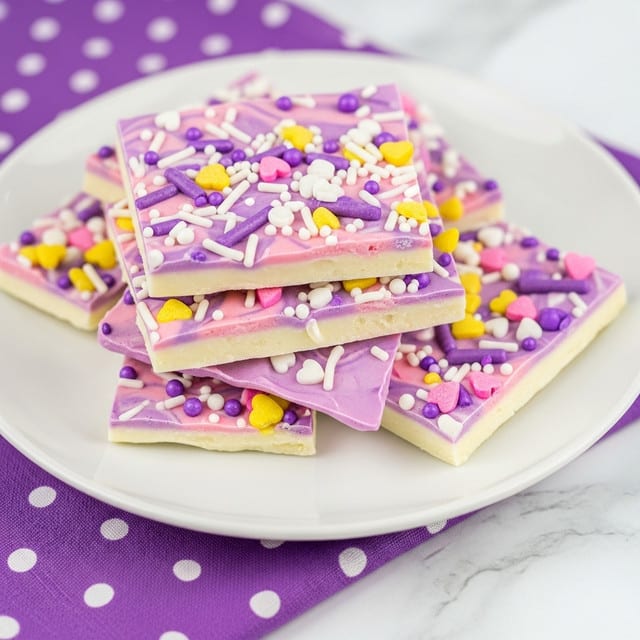 A white plate sits on a white marbled surface with several pieces of colorful bark candy stacked on it. The bark has two layers; the bottom layer is mostly white and the top layer is a mix of light pink and purple swirls. The entire bark is covered with small sprinkles in white, yellow, purple, and pink, including heart shapes, dots, and small sticks. The plate is partly resting on a purple cloth with white polka dots. photo taken with an iphone --ar 4:5 --v 7