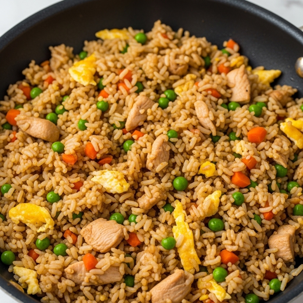 A close-up image of a cooked fried rice dish showing one main layer filled with cooked long-grain rice mixed with small green peas, diced orange carrots, scrambled egg bits, and pieces of light brown chicken. The rice is seasoned with soy sauce giving it a light brown color that covers the grains evenly. The dish is inside a cooking pan with a dark non-stick surface, and the background shows a white marbled texture. photo taken with an iphone --ar 4:5 --v 7