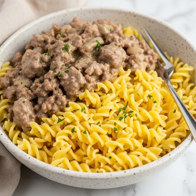 A close-up of a bowl of creamy ground beef pasta with spiral noodles, showing one layer of curled yellow pasta mixed fully with small bits of brown cooked ground beef, all coated in a smooth light brown sauce. The pasta and beef mix fills the bowl almost to the top, with a slight sheen from the sauce. The bowl is a speckled white ceramic, placed on a white marbled surface, and a fork rests on the right side inside the bowl. Photo taken with an iphone --ar 4:5 --v 7