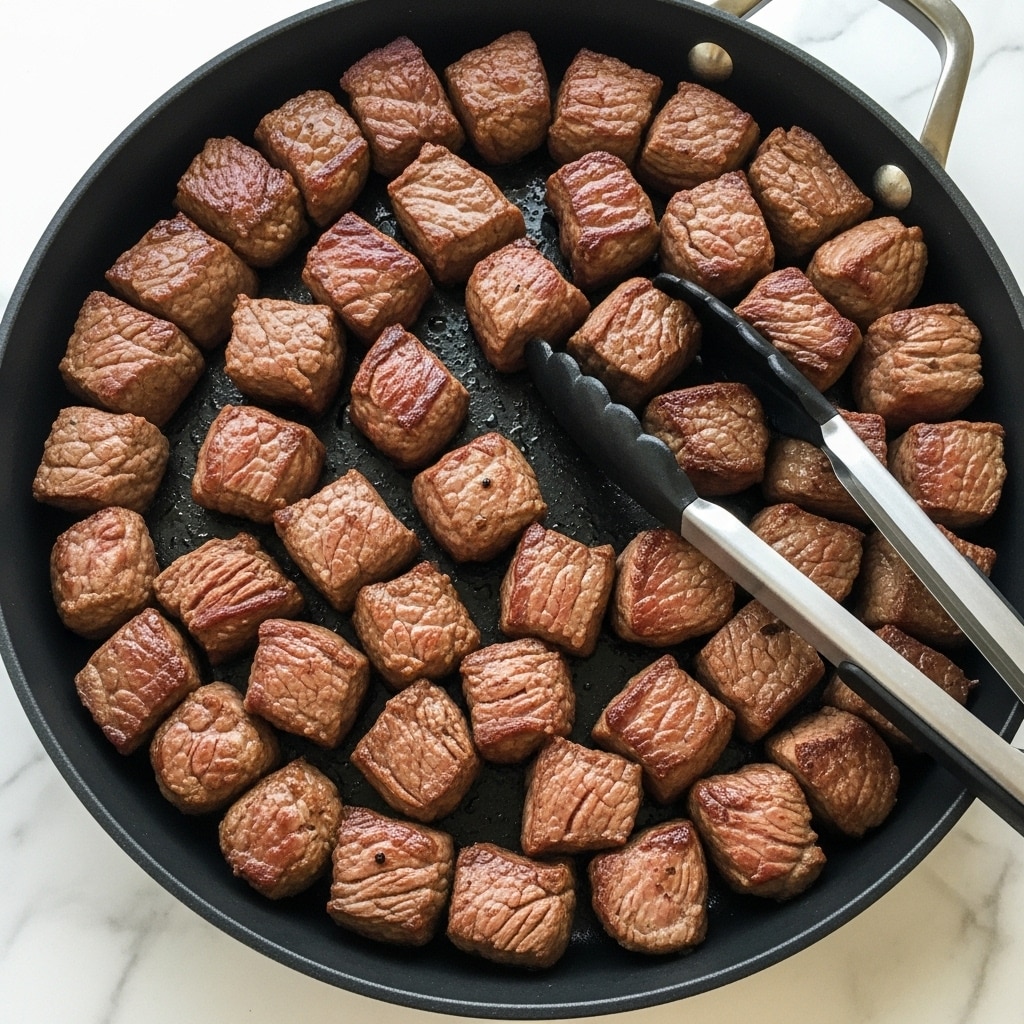The image shows many small, browned beef cubes cooked inside a black cast iron skillet with a light green handle, placed on a white marbled surface. The beef pieces are unevenly cut into bite-sized chunks with a slightly crispy outside and moist interior, scattered over the entire pan. A pair of metal tongs with black silicone tips rest among the beef pieces, with one tip partially touching the meat and the other resting on the skillet. The skillet edges show some glistening oil spots, adding a shiny texture to the cooking surface. Photo taken with an iphone --ar 4:5 --v 7