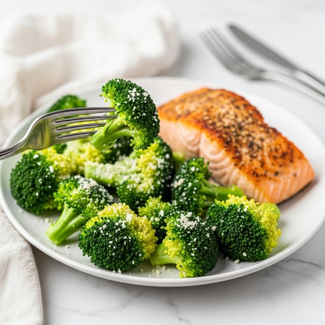 A white plate holds a serving of bright green broccoli florets sprinkled with white grated cheese, with a piece of cooked salmon that has a golden-brown, slightly crispy surface placed at the back right side of the plate. A silver fork lifts one broccoli floret prominently in the foreground. The plate is set on a white marbled surface, with another fork and a soft white cloth napkin nearby. Photo taken with an iphone --ar 4:5 --v 7