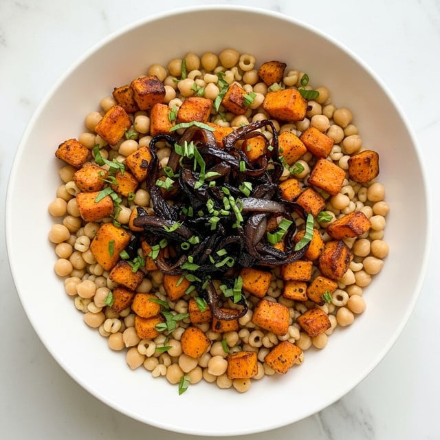 A white bowl holds a colorful dish made of small round pasta pearls as the bottom layer mixed evenly with light brown chickpeas and golden roasted cubed sweet potatoes. On top, there are scattered dark caramelized onion slices and small green herb pieces, creating a mix of textures and colors. The dish sits on a white marbled surface. photo taken with an iphone --ar 4:5 --v 7