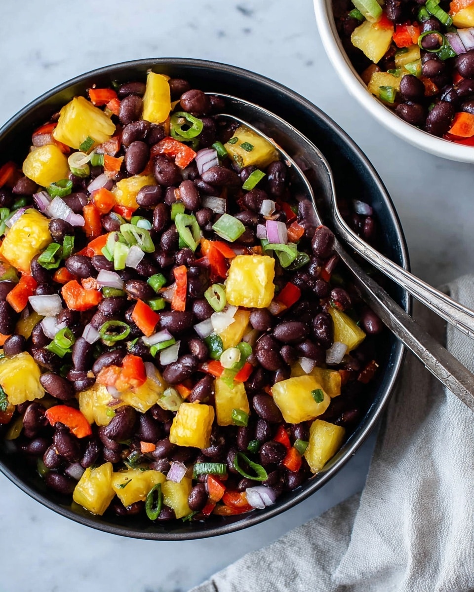 A close-up view of a black bowl filled with a colorful black bean salad, showing three main layers: the bottom layer of dark purple black beans, scattered thick chunks of bright yellow pineapple pieces, and pieces of red bell peppers on top mixed with small rings of white onions and bits of green onions, all evenly mixed. Two silver spoons rest inside the bowl horizontally, partially covered by the salad. To the top right, a white bowl with similar contents is partially visible on a white marbled surface, next to a light gray cloth. The lighting is bright and natural, showing a fresh and vibrant dish. Photo taken with an iphone --ar 4:5 --v 7