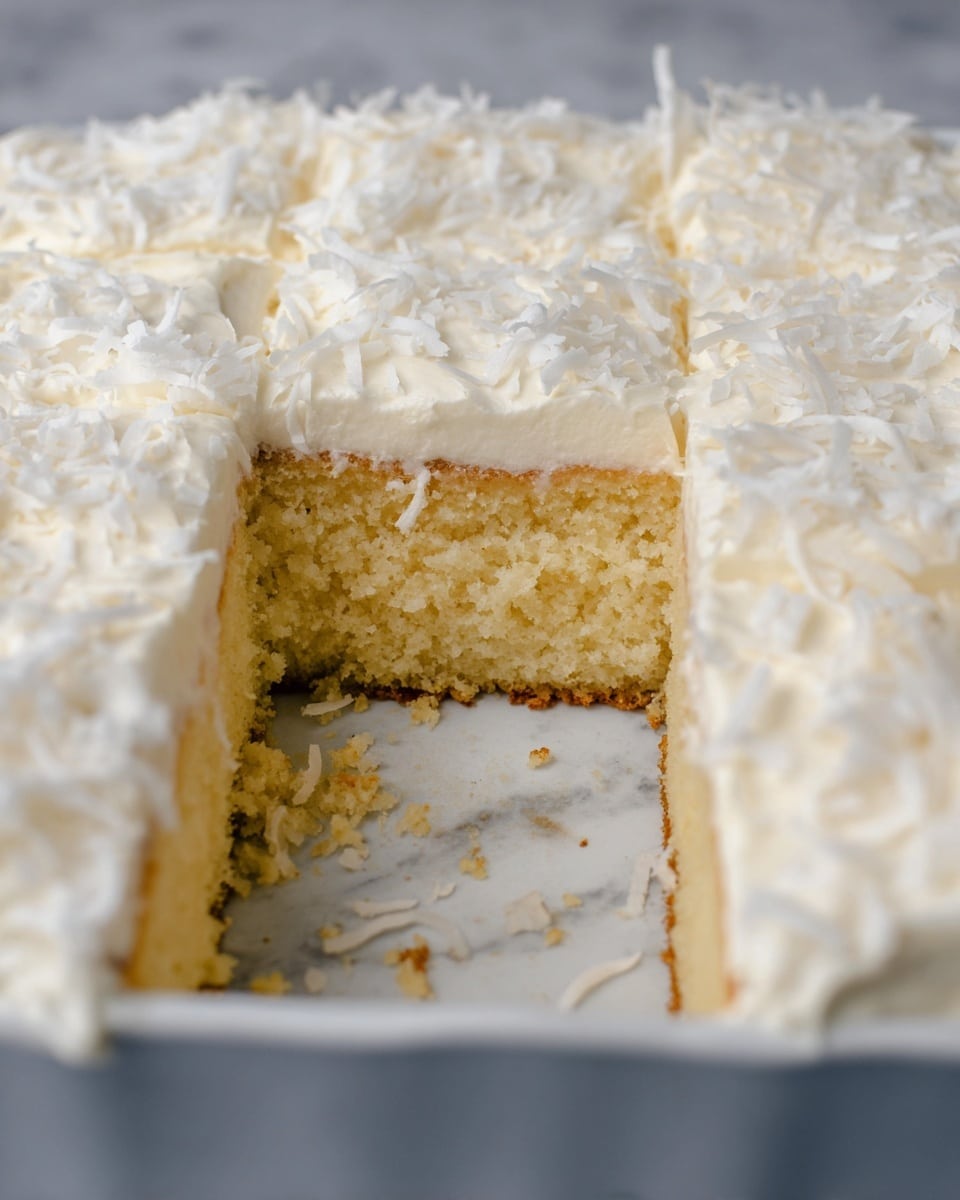 A dessert with two visible layers sits on white plates and in a clear glass baking dish on a blue and white checkered cloth over a white marbled surface. The bottom layer is a light yellow cake with a soft texture. The top layer is thick white frosting covered with white coconut flakes. The piece on the plate in front is square and has a silver fork resting next to it. The baking dish also holds multiple square pieces, with one piece missing from the corner. photo taken with an iphone --ar 4:5 --v 7