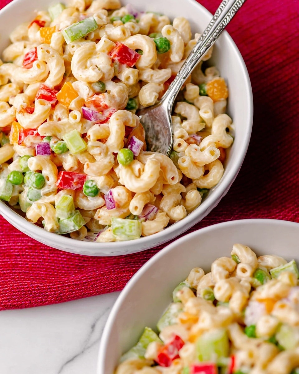 A close-up of a white bowl filled with colorful macaroni salad placed on a white marbled textured surface with a red cloth underneath. The salad has two visible layers: the first layer consists of creamy elbow-shaped macaroni pasta coated in a light dressing, and the second layer has evenly mixed, diced pieces of green celery, red bell pepper, orange cheddar cheese cubes, green peas, and small bits of red onion scattered throughout. A silver spoon is partially inside the bowl, slightly digging into the salad. In the lower part of the image, part of another white bowl filled with the same salad is visible. photo taken with an iphone --ar 4:5 --v 7