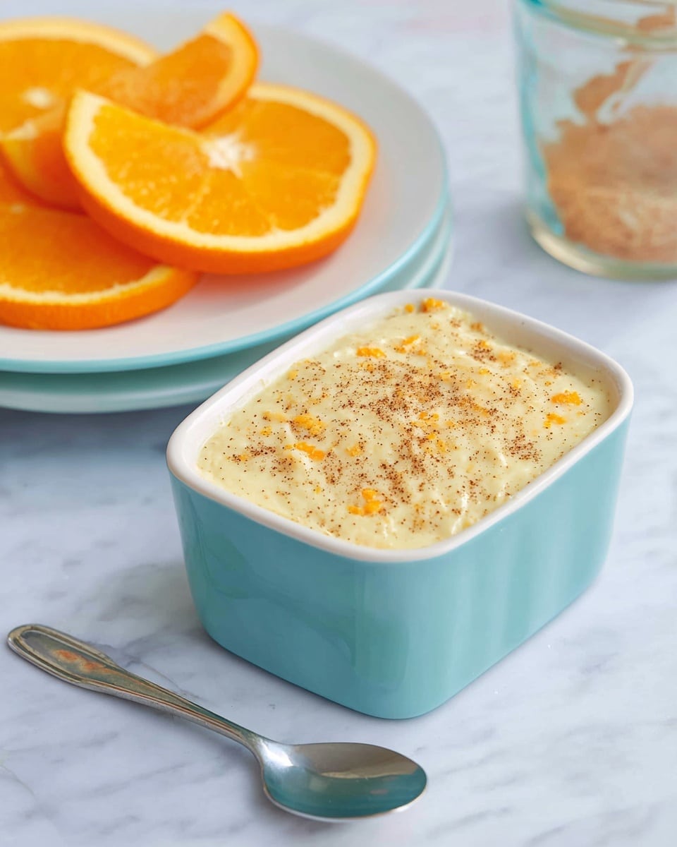 A small light blue rectangular bowl filled with a creamy dessert that has a pale yellow color with small orange bits mixed inside and a light sprinkle of brown spice on top. Behind the bowl, on a white plate, there are three bright orange slices with juicy texture. A silver spoon is placed in front of the bowl on a white marbled surface, creating a clean and bright setting. photo taken with an iphone --ar 4:5 --v 7