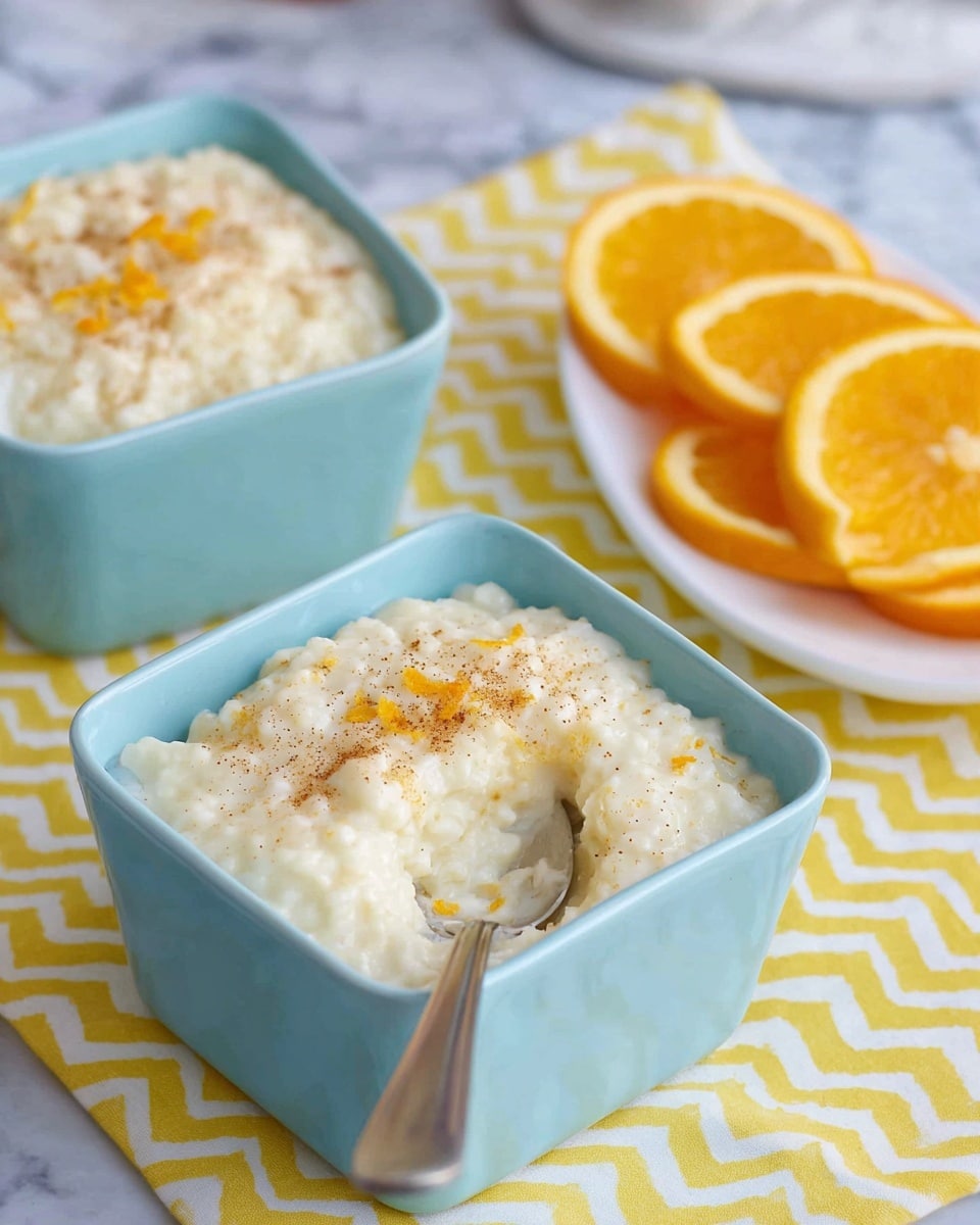 The image shows two light blue square bowls filled with creamy rice pudding. The pudding has a smooth, thick texture with a pale cream color, lightly sprinkled with brown cinnamon and small orange zest bits on top. One bowl in the front has a silver spoon scooping out some pudding, revealing its slightly chunky, soft rice texture inside. Behind the bowls, there is a white plate holding two orange slices, placed on a yellow zigzag patterned cloth over a white marbled surface. The photo taken with an iphone --ar 4:5 --v 7