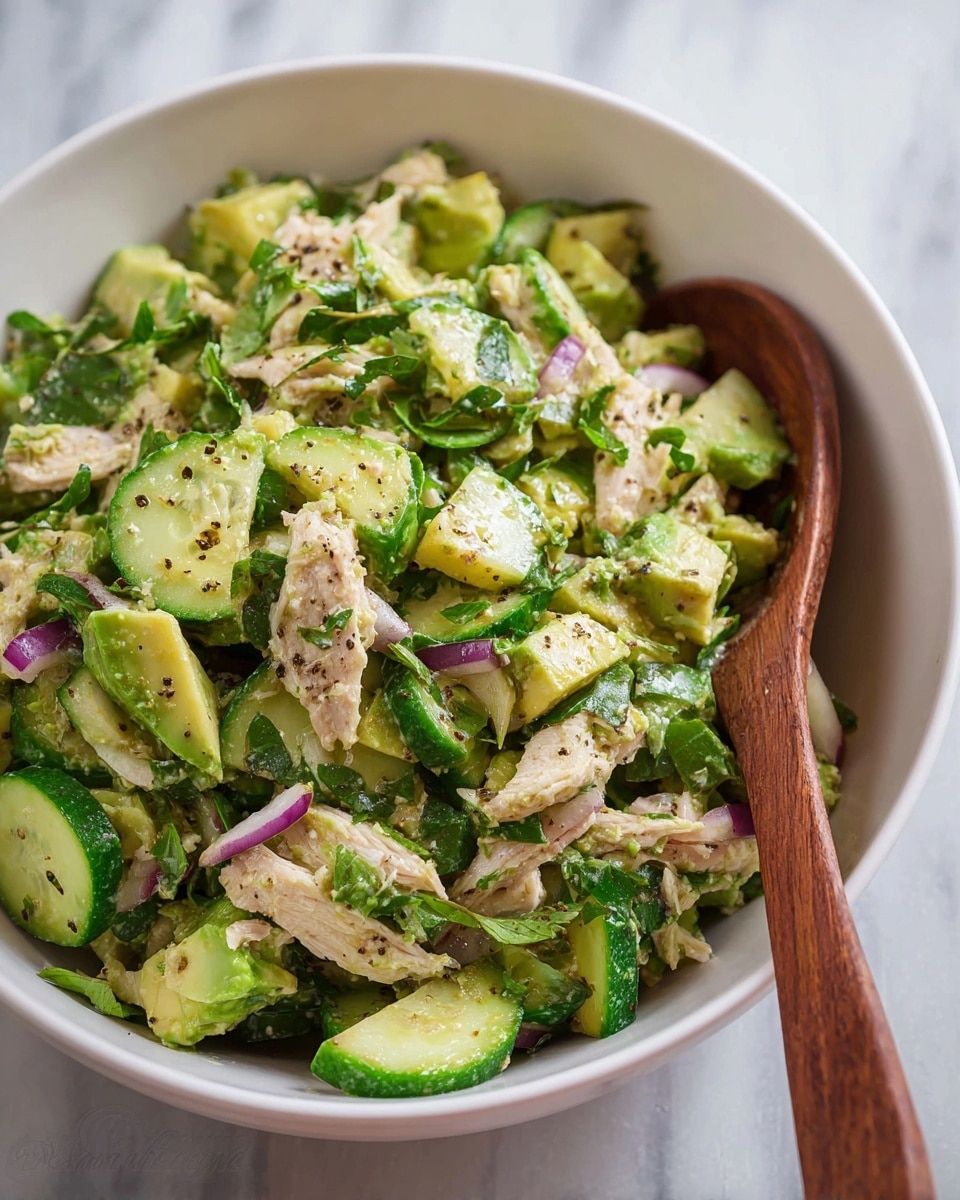In a white bowl, the dish has four main layers divided into sections: light tan shredded chicken seasoned with black pepper on the top right; bright green chopped cilantro in the center; smooth, light green diced avocado with a hint of olive oil and black pepper on the bottom left; thin slices of purple onion on the far left; and fresh, green cucumber slices sprinkled lightly with black pepper on the top left. The bowl sits on a white marbled surface with a blurred background that includes half a yellow lemon and some wooden utensils nearby. Photo taken with an iphone --ar 4:5 --v 7