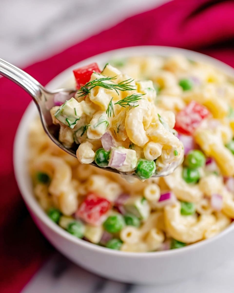 A close-up of a white bowl filled with creamy macaroni salad, featuring three layers packed together: the first layer is pale yellow elbow macaroni coated in a smooth dressing, the second layer includes bright green peas and finely chopped red onion pieces scattered throughout, and the third layer has small cubes of light yellow cheese and red bell pepper, all garnished with tiny green dill sprigs. A metal spoon holds a portion of the salad above the bowl, showing the mix’s soft and fresh textures, with a white marbled surface underneath and a blurred red cloth in the background. Photo taken with an iphone --ar 4:5 --v 7