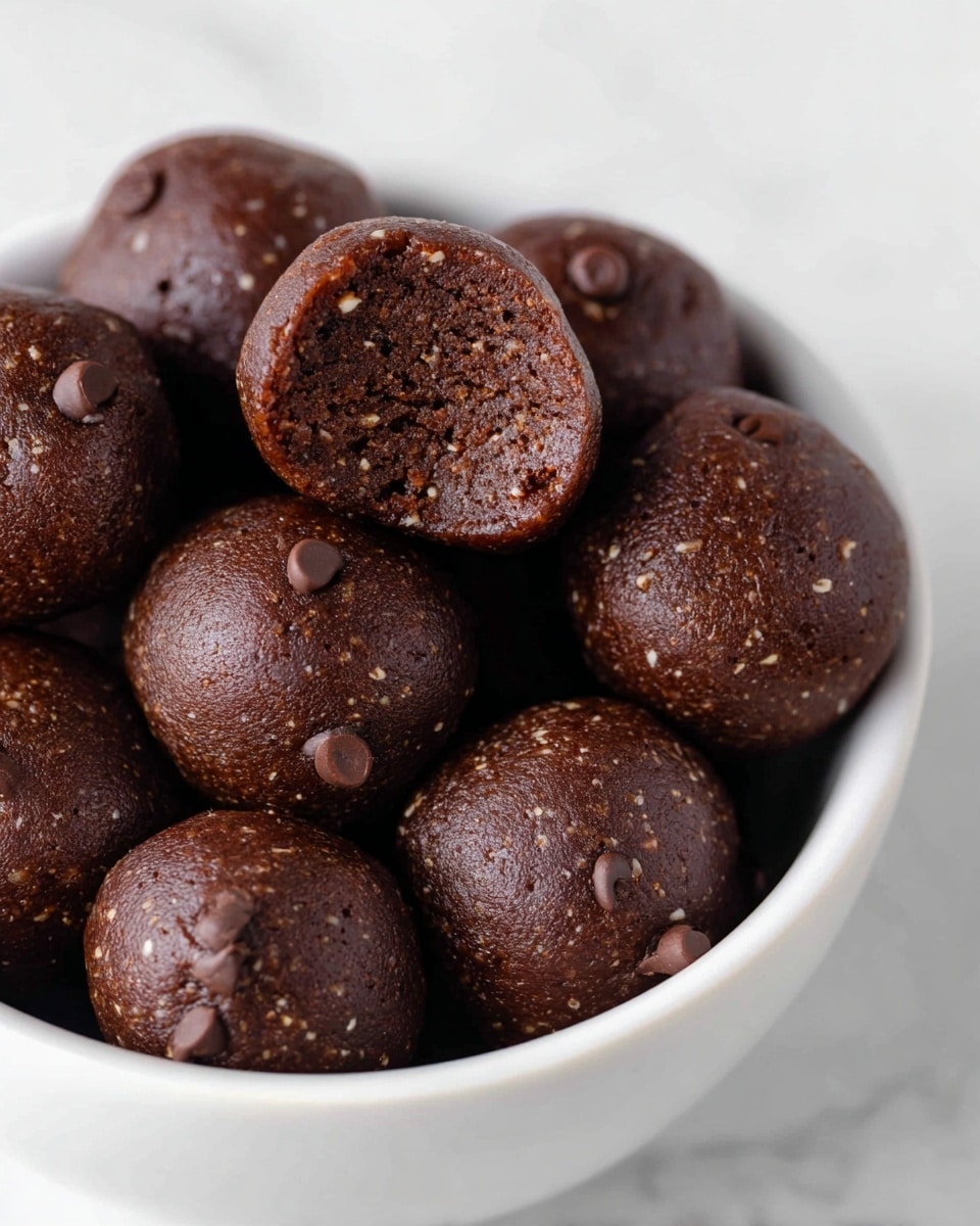 A close-up view of a white square bowl filled with round, dark brown energy balls dotted with small chocolate chips. Each ball has a slightly textured surface and is tightly packed inside the bowl, showing a rough but moist exterior. A second white bowl with more energy balls is blurred in the background, placed on a white marbled surface. The overall look is simple and clean with a focus on the rich chocolatey color and texture of the balls. Photo taken with an iphone --ar 4:5 --v 7