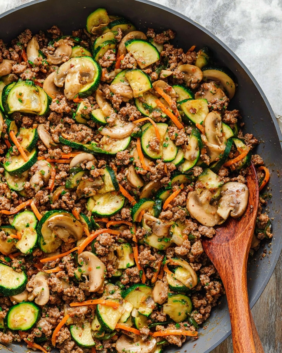 A close-up of a large grey pan filled with cooked ground meat mixed with sautéed green zucchini slices, light brown sliced mushrooms, and thin orange carrot strips, all seasoned and lightly browned. A wooden spoon rests inside the pan on the right side, partially covered by the food. The background is a white marbled texture. The colors are warm and natural, showing a well-cooked mixture of vegetables and meat. photo taken with an iphone --ar 4:5 --v 7