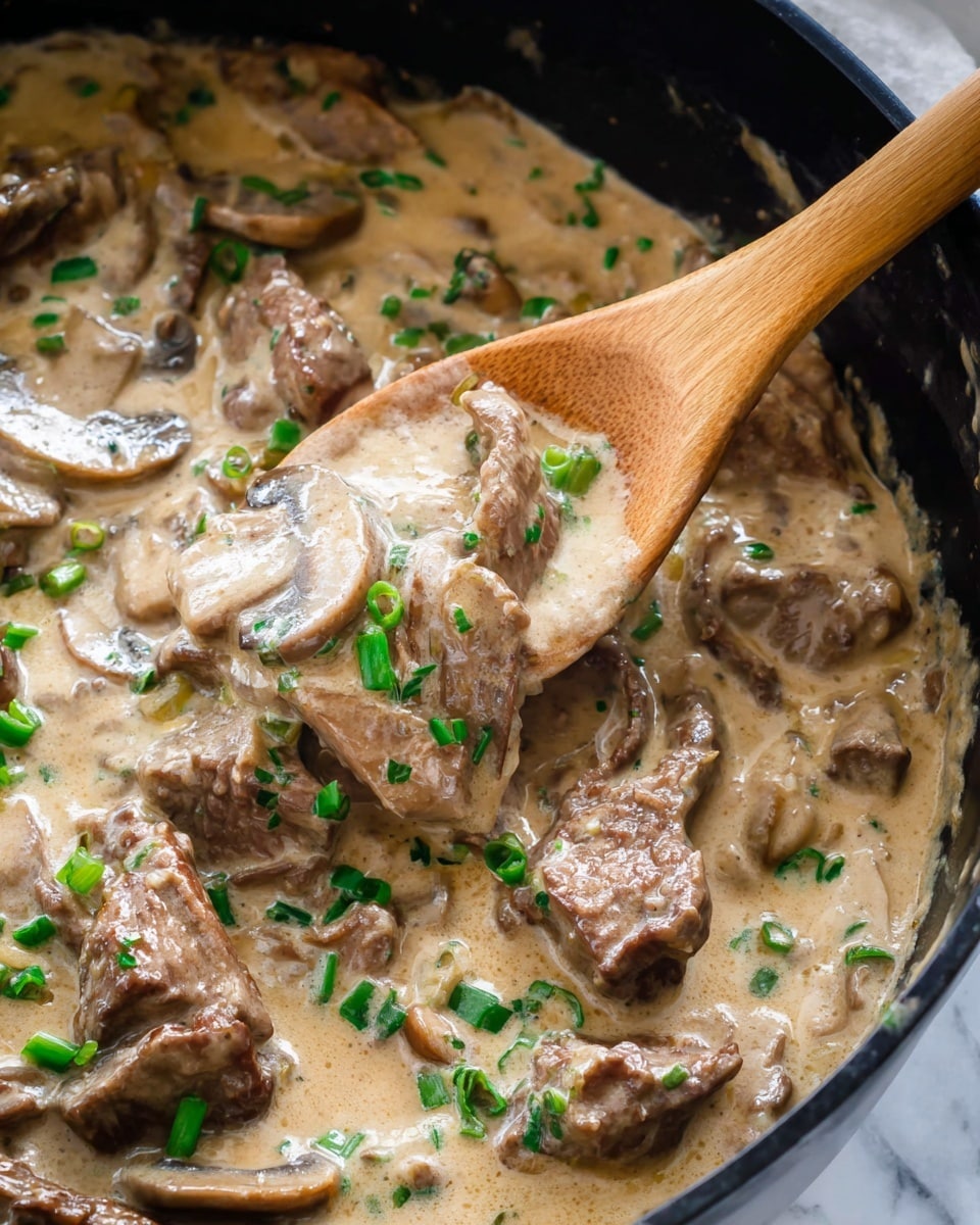 A close-up view of a dark pan filled with creamy light brown mushroom sauce mixed with slices of light and dark brown mushrooms and pieces of browned meat. The sauce has a smooth texture and is sprinkled with small bright green chopped chives on top. On the right side of the pan, a wooden spoon with a pale handle is resting in the sauce. The pan is placed on a white marbled surface with a red and white checkered cloth nearby. Photo taken with an iphone --ar 4:5 --v 7