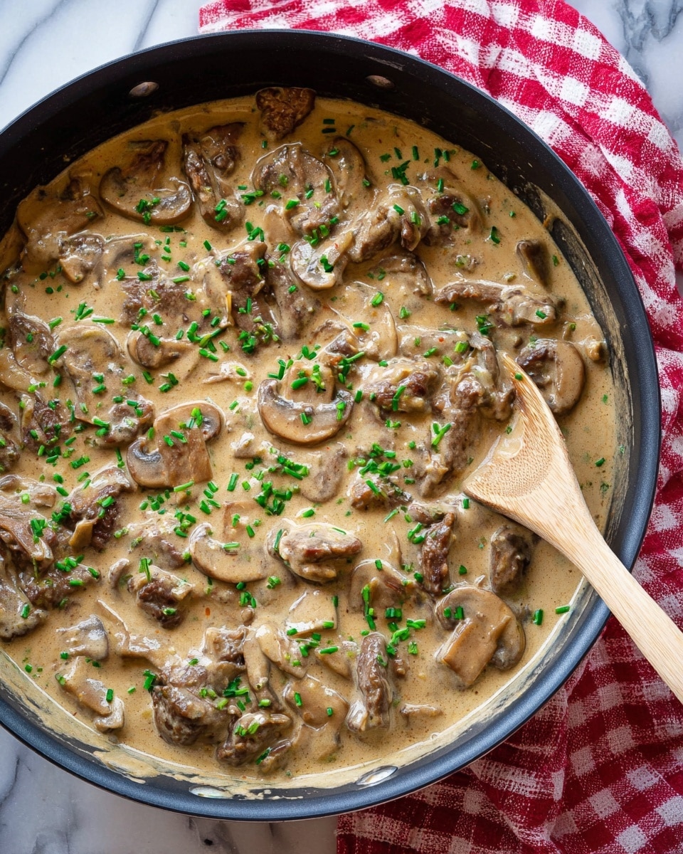 This is a close-up view of a creamy mushroom and beef dish in a black pan, with a wooden spoon lifting a portion. The dish has several layers: thick brown pieces of cooked beef, mixed with soft, light brown mushroom slices covered in a pale beige creamy sauce that looks smooth and rich. Small, bright green chopped scallions are sprinkled on top, adding a fresh color contrast. The textures show the softness of the mushrooms, the slight firmness of the beef, and the creamy sauce surrounding all pieces. The background is a white marbled texture. photo taken with an iphone --ar 4:5 --v 7