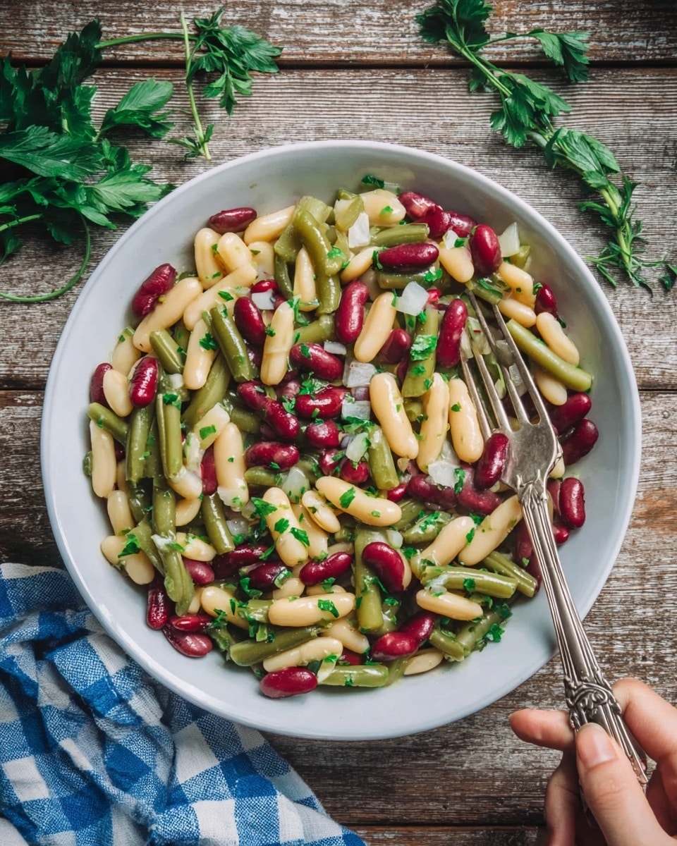 The image shows a close-up of a white shallow bowl filled with a colorful bean salad. The salad has three layers: the bottom layer is mostly pale yellowish beans, mixed with a smaller amount of red kidney beans and green beans all evenly spread. On top, there are small pieces of chopped white onion and bright green chopped parsley scattered over the beans. A woman's hand holds a silver fork resting inside the bowl on the right side. The bowl is placed on a rustic wooden table with a blue and white checked cloth to the left and some fresh herbs around it. The photo is taken with an iphone --ar 4:5 --v 7