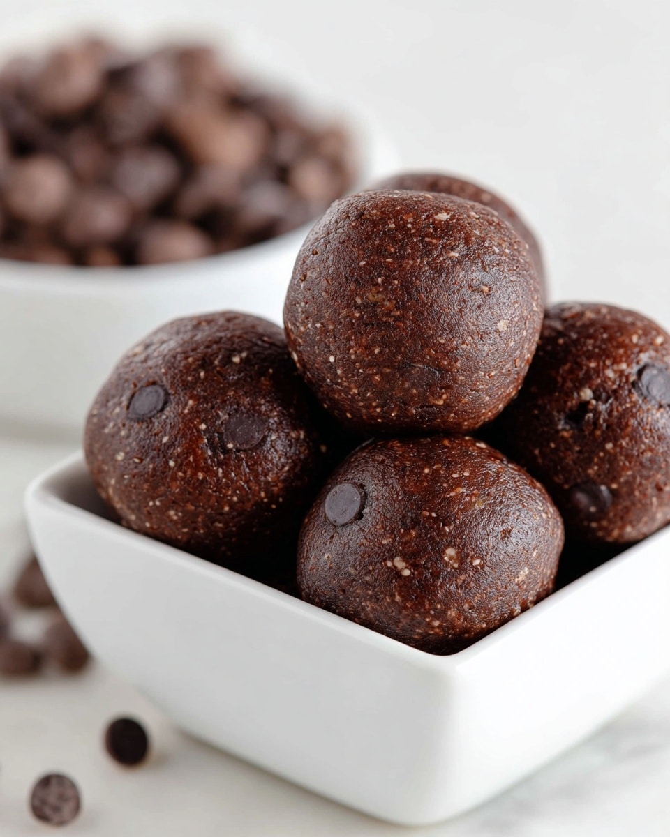 A close-up image of a white bowl filled with round, dark brown chocolate energy balls, each with a smooth, slightly shiny texture and small chocolate chips embedded on the surface; one energy ball is slightly bitten, showing a dense, moist, and textured inside with tiny bits of nuts or oats distributed throughout. The bowl sits on a white marbled texture. photo taken with an iphone --ar 4:5 --v 7