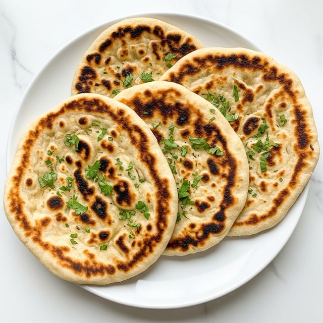 Three naan flatbreads are stacked slightly overlapping on a white plate, each showing a golden brown, charred texture with uneven darker spots. The flatbreads are sprinkled with small pieces of green herbs scattered on top, adding a fresh contrast to the warm, toasted surface. The plate rests on a white marbled surface, enhancing the clean and simple presentation. photo taken with an iphone --ar 4:5 --v 7