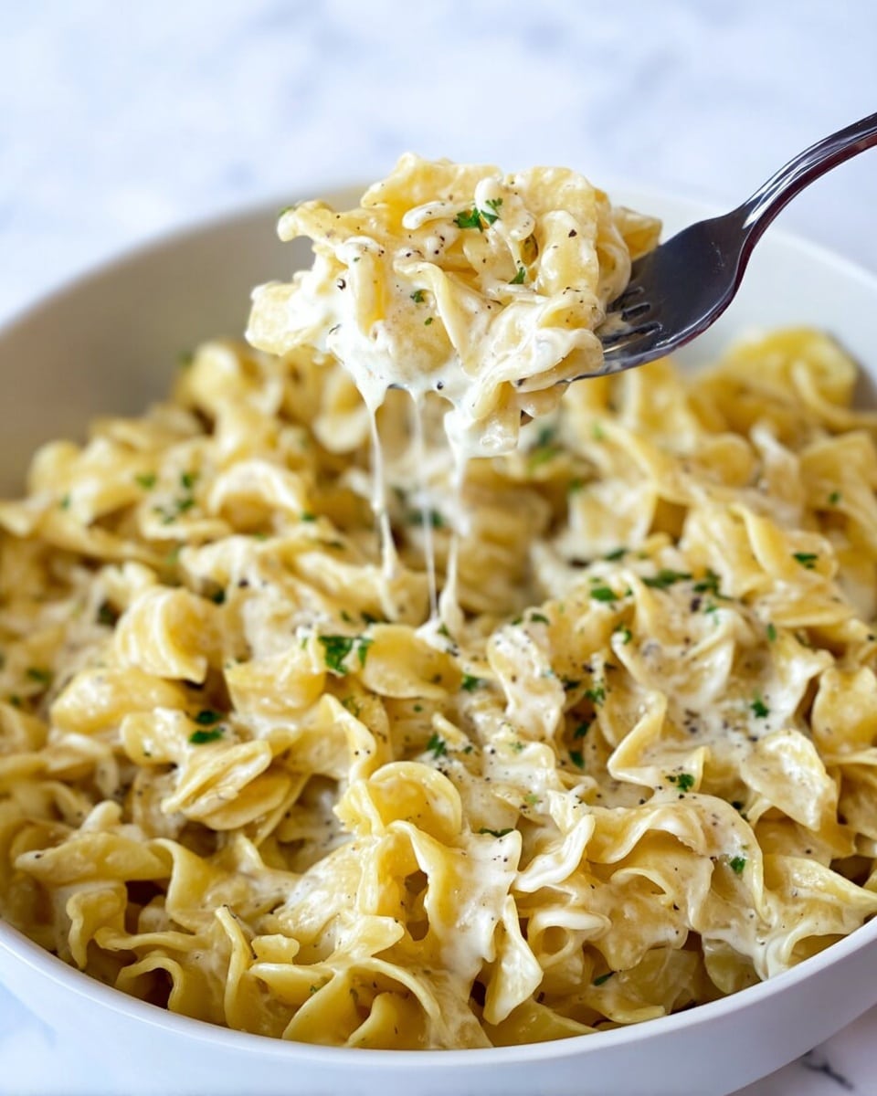 Two white bowls filled with creamy egg noodle pasta sit on a white marbled surface. Each bowl has crinkled yellow pasta with a smooth, thick white sauce coating the noodles. Small white curds, likely cottage cheese, are scattered evenly throughout the pasta layers. The top of the pasta is sprinkled with finely chopped green herbs and cracked black pepper, adding contrast to the creamy yellow noodles. In the background, a dark turquoise pot containing more pasta is partially visible, along with a gray textured cloth and a silver fork resting beside the bowls. The scene is bright and focused, showcasing the pasta texture and toppings clearly, photo taken with an iphone --ar 4:5 --v 7