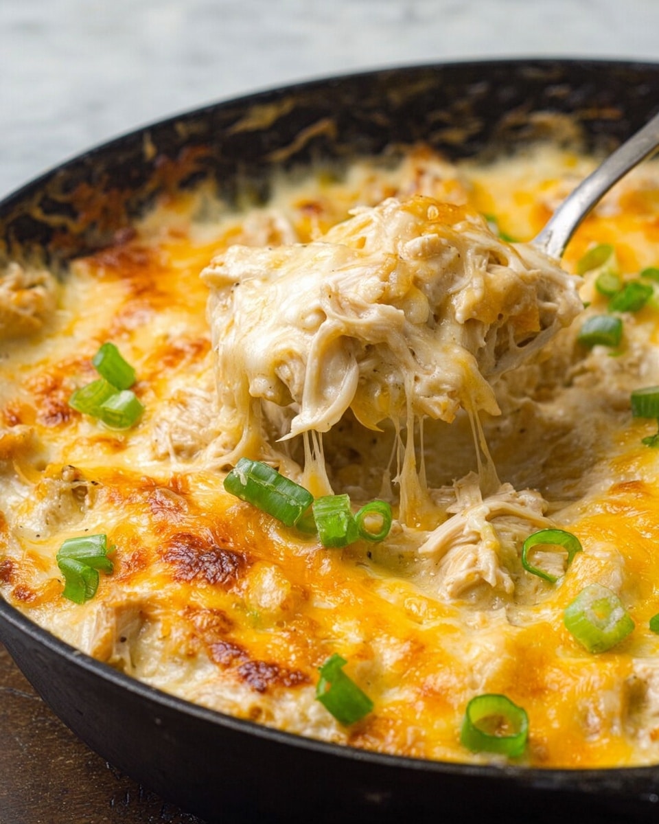 A close-up view of a baked dish in a black cast iron pan shows a creamy base layer mixed with bits of green, likely green onions, and chunks of light-colored chicken. On top, a golden layer of melted cheddar cheese covers most of the surface, with some browned spots indicating it is baked. Scattered pieces of chopped green onions sit on the cheese, adding bright green contrast. The creamy sauce around the edges looks smooth and rich, blending with the cheese in a bubbly texture. photo taken with an iphone --ar 4:5 --v 7