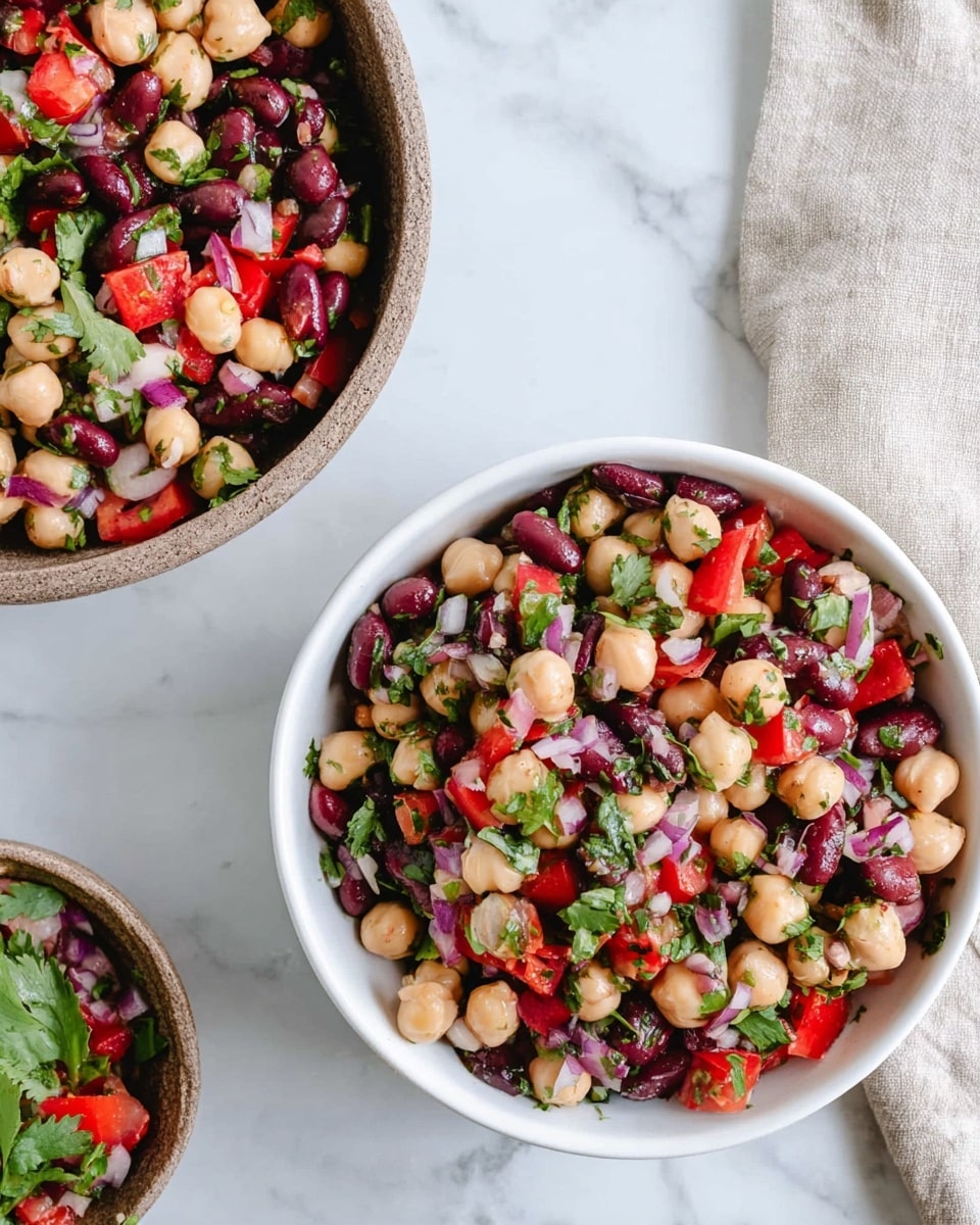The image shows two white bowls filled with colorful bean salad placed on a white marbled surface. Each bowl holds a mix of three types of beans—light beige chickpeas, dark red kidney beans, and small black beans—mixed with bright red bell pepper pieces and finely chopped purple onions. Fresh green herbs, likely cilantro, are sprinkled throughout the salad adding bursts of green color. The larger bowl sits near the top right of the image, with a part of a beige cloth napkin visible nearby, and the smaller bowl is placed lower left, slightly overlapping the napkin's edge. The textures show the smooth roundness of the beans and the fresh crispness of the peppers and herbs. Photo taken with an iphone --ar 4:5 --v 7
